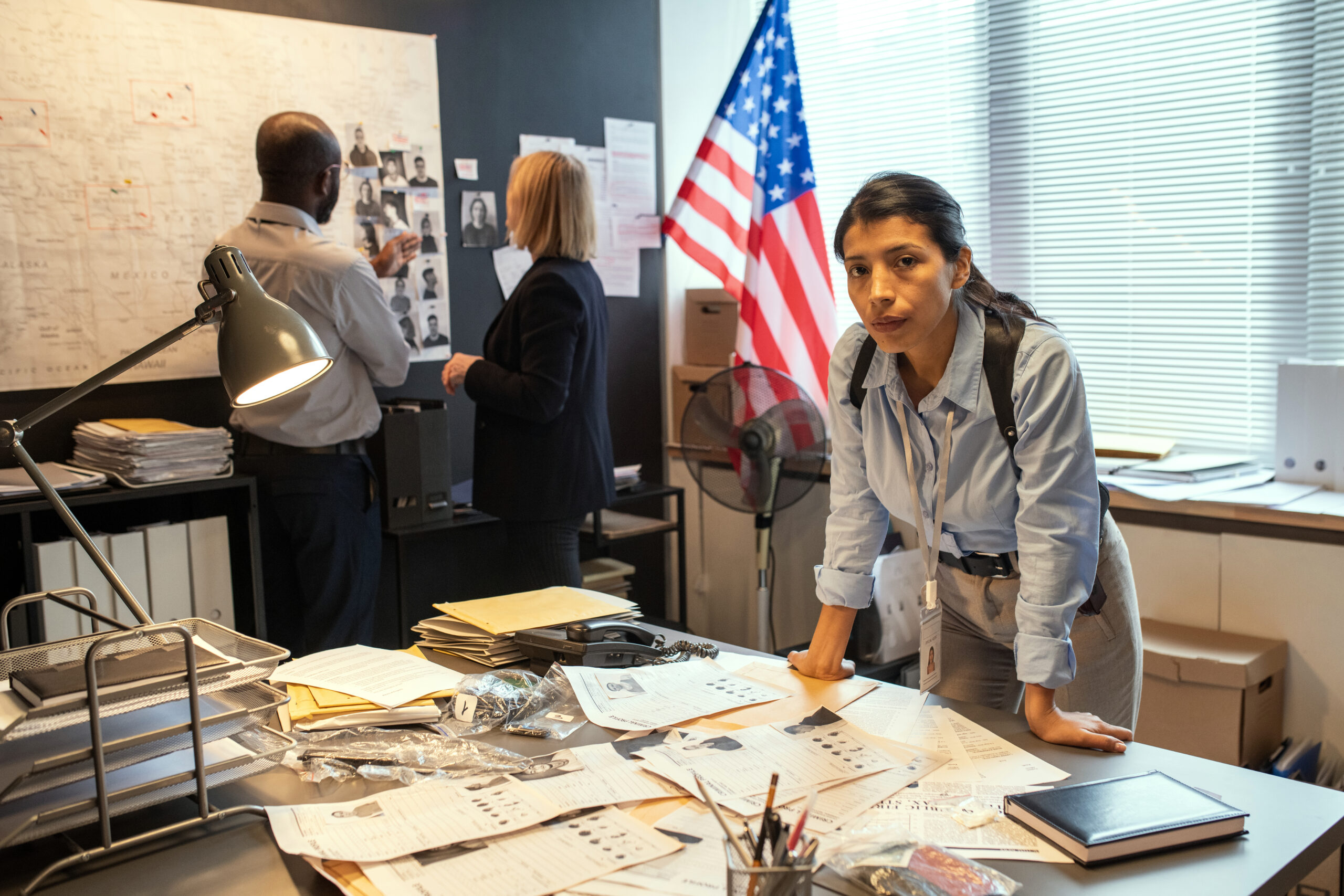 A photo of a young Hispanic female detective standing over a desk covered with papers. Behind her, two other detectives look at a wall covered in photographs and clues. 