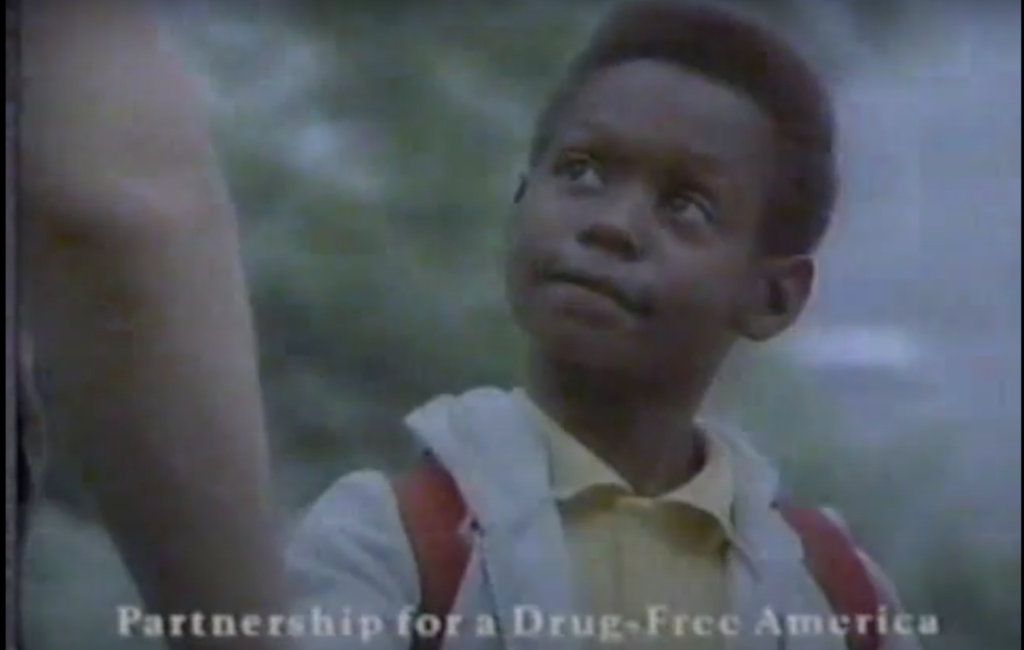 Screenshot of a 1990 public service announcement from the partnership for a drug-free America, showing a iblack child wearing a backpack looking up at an adult who is just off-screen. 